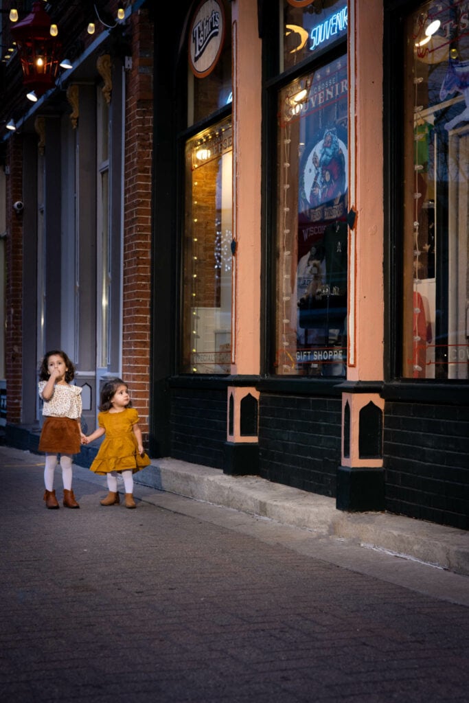 Two young girls in yellow dresses stand on a European street at dusk, illuminated by storefront lights