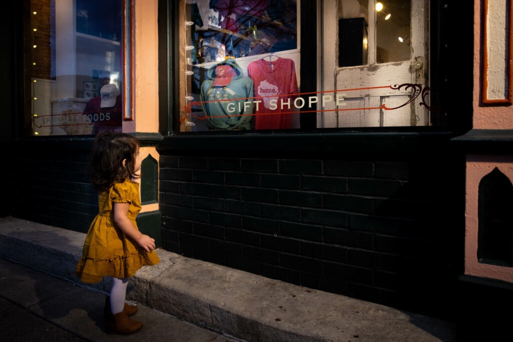 Child in yellow dress admiring gift shop window display at dusk, warm street lighting