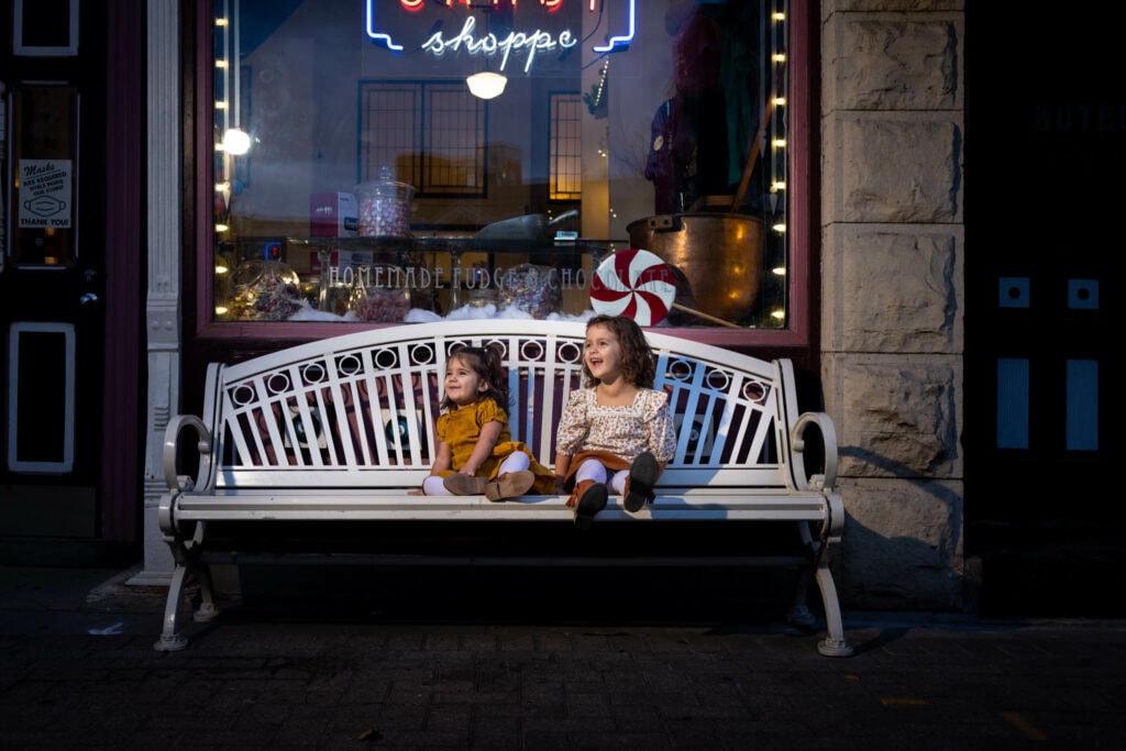 Two young children sitting on an illuminated bench in front of a holiday-decorated storefront at night