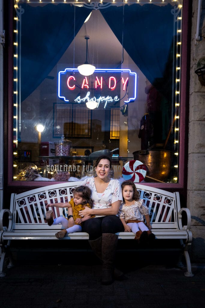 Woman with two young children sitting on bench beneath glowing candy shop neon sign at night