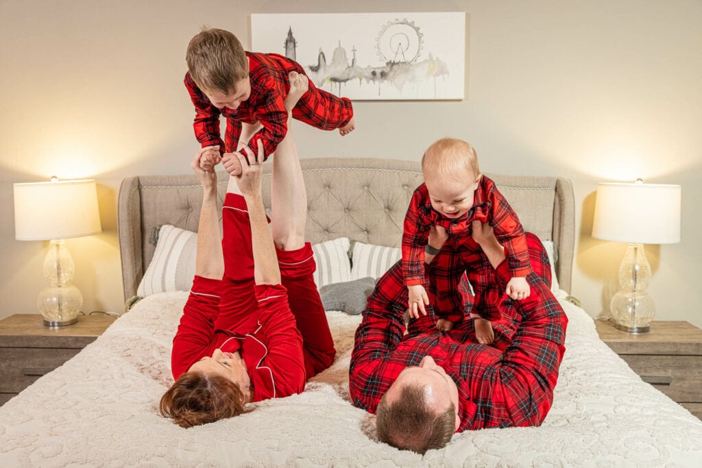 Young children in red plaid pajamas play on a bed, one lifted in the air, during a family portrait session