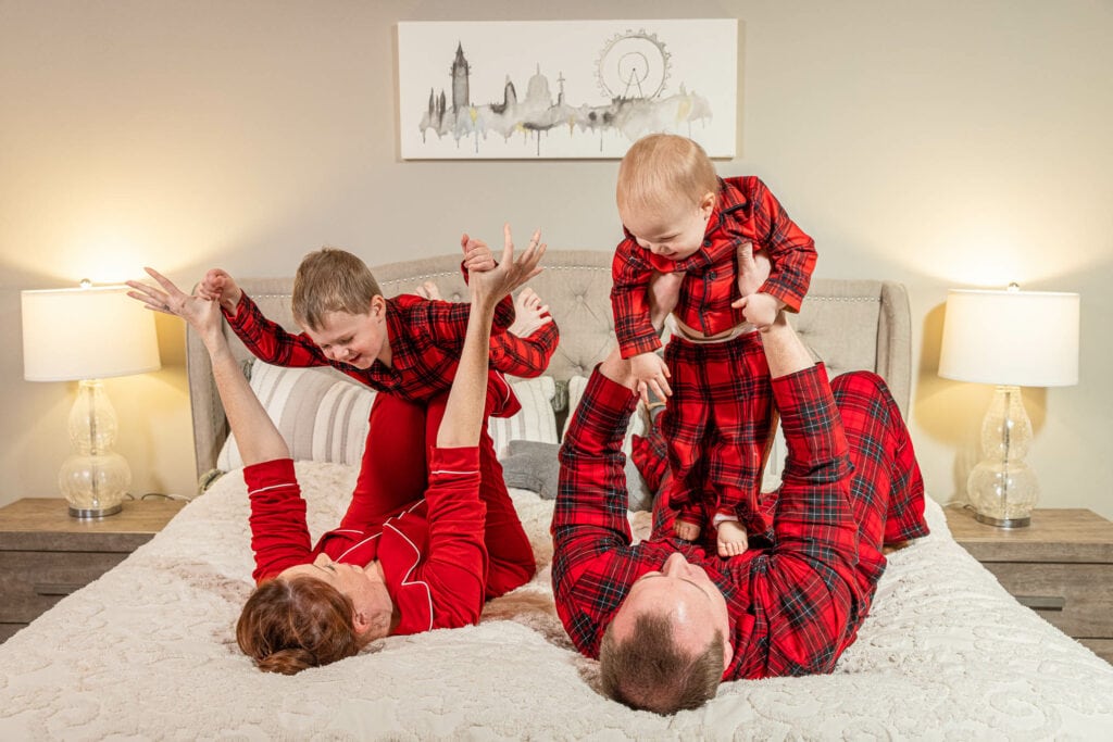 Family of four in matching red plaid pajamas playing on bed, warm bedroom lighting, holiday portrait