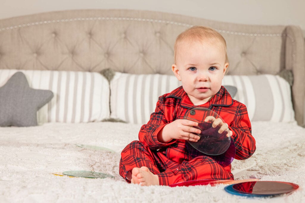 Toddler in red plaid pajamas sitting on bed, holding smartphone and smiling at camera