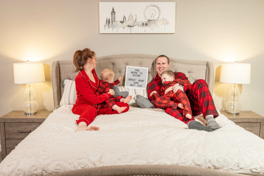 Family of four in matching red pajamas on bed holding Christmas card, warm bedroom lighting
