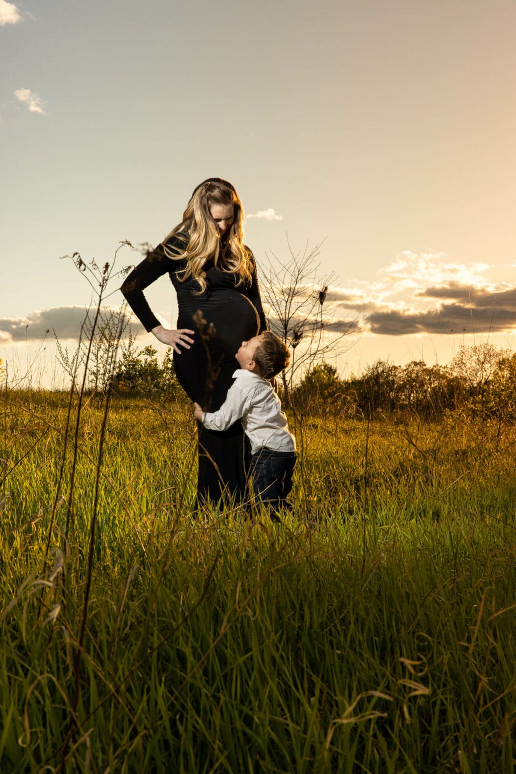 A pregnant mother stands in a golden-hour field as her young son hugs her legs and looks up at her