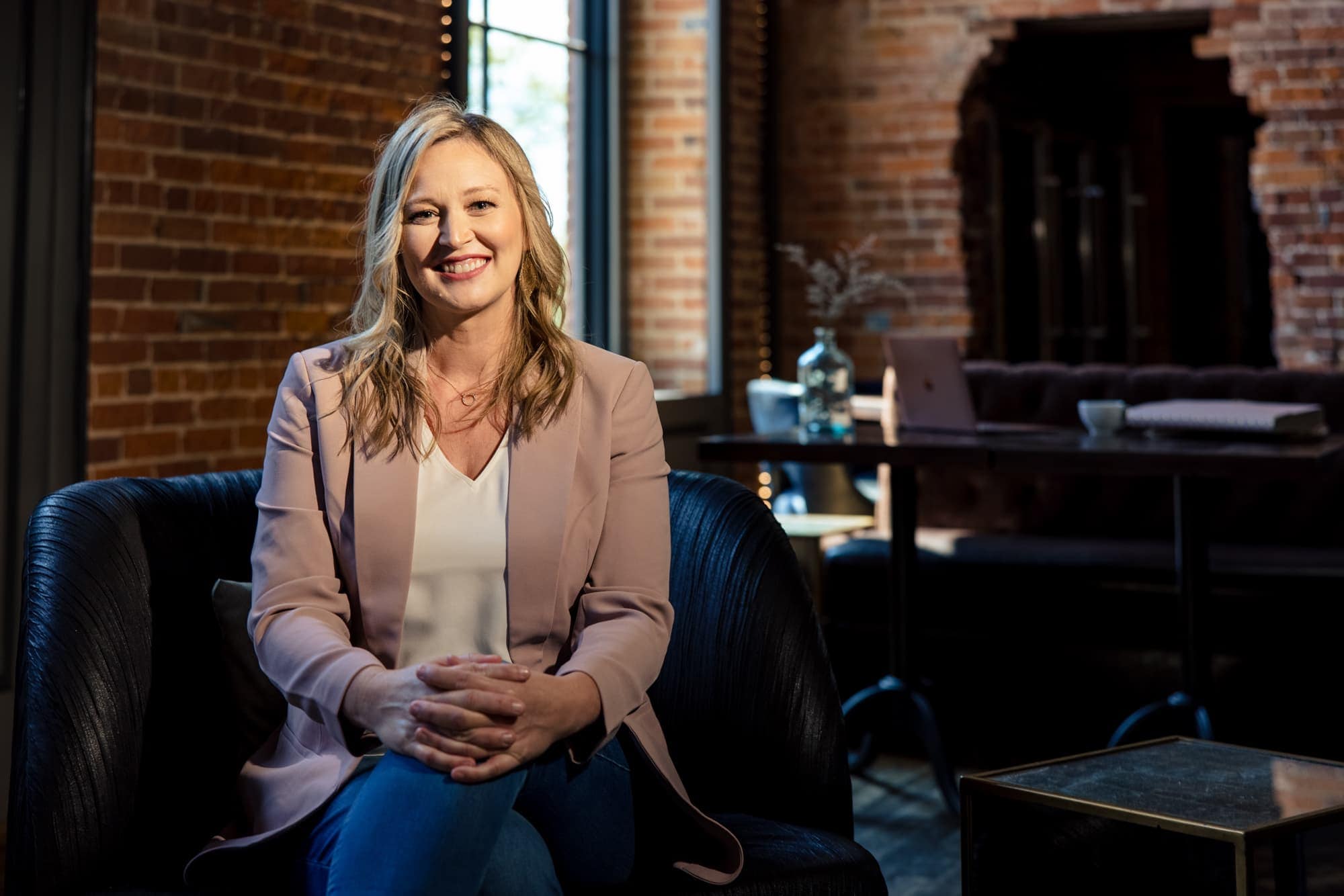Seated in a brick-walled studio in a taupe blazer