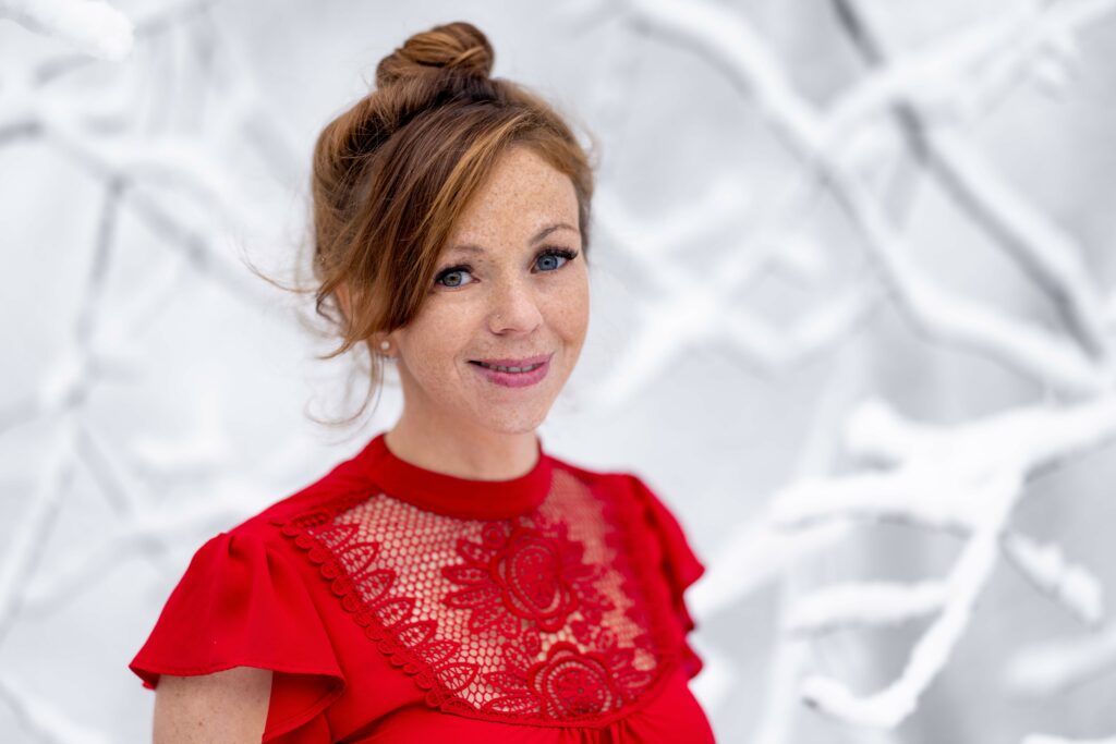 Young woman in red shirt with professional makeup, hair in top bun, smiling against white textured backdrop