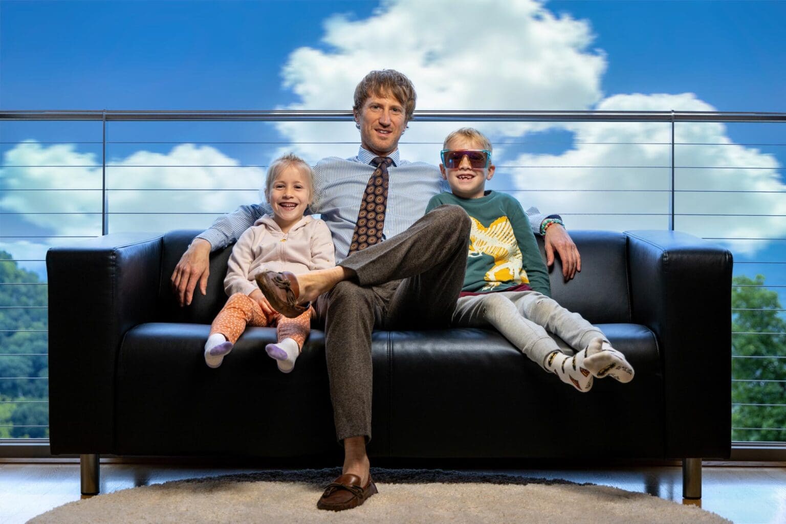 Father with two children on a black sofa by a sky-view window
