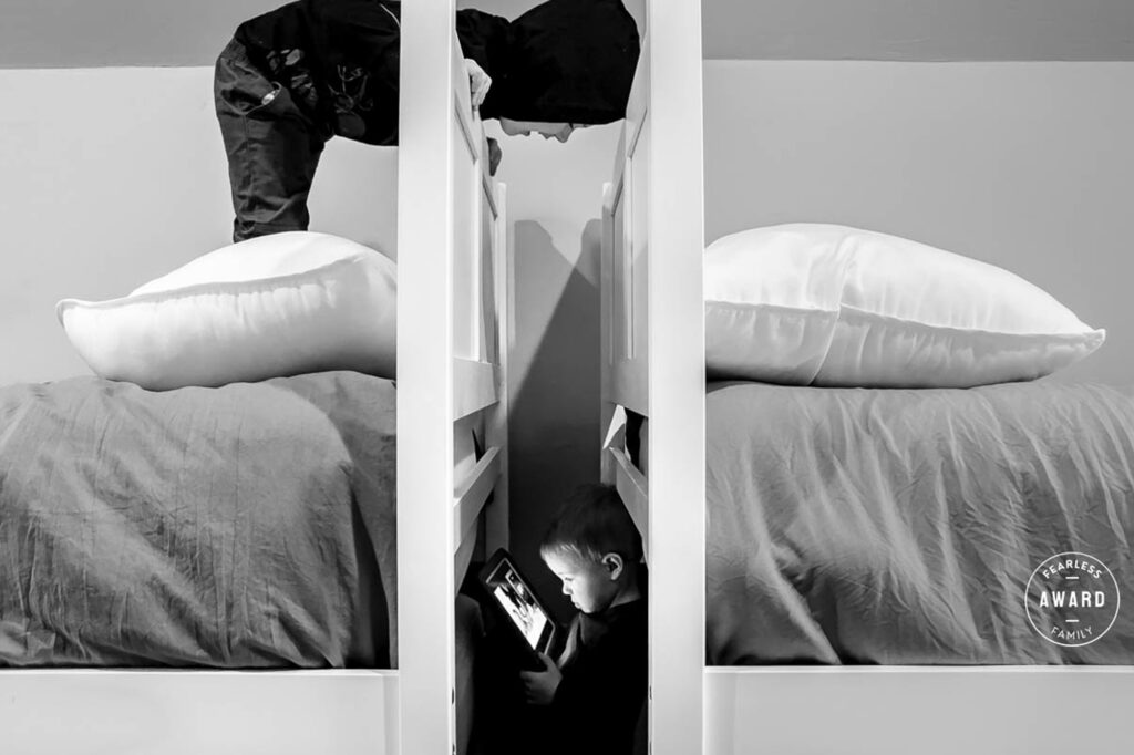 Black and white portrait of child sitting between mirrored panels, holding phone, creating reflection effect on bed with pillows