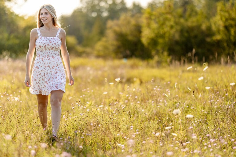 Blonde woman in floral dress walking through sunlit meadow with wildflowers and trees