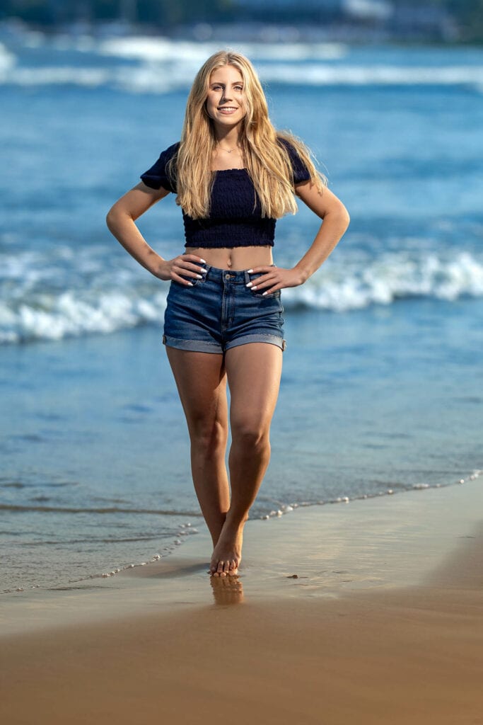Blonde teenage girl in black crop top and denim shorts standing on beach, ocean waves and sand visible behind her