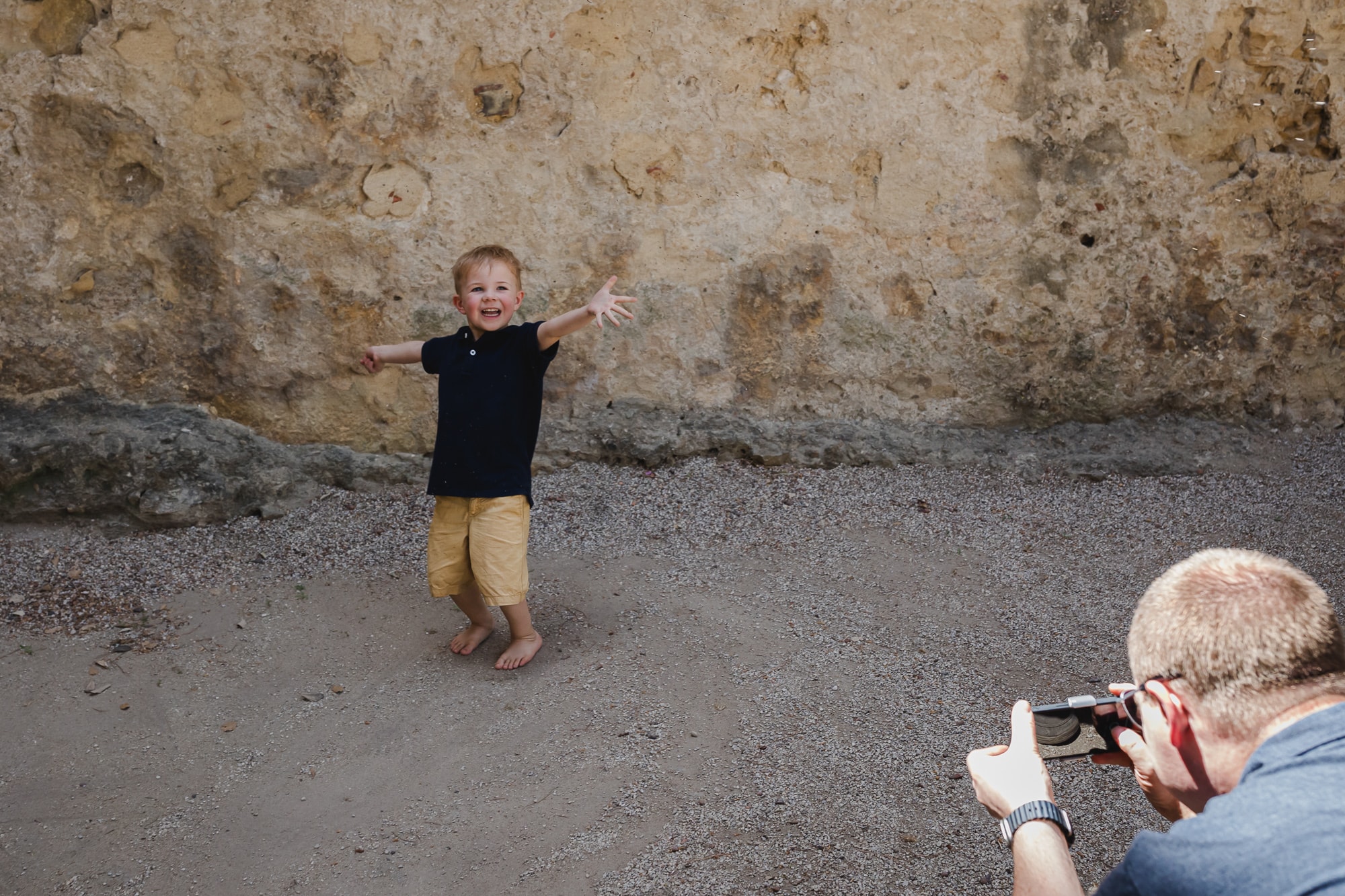Toddler with arms spread wide smiling at camera while parent photographs in Lisbon