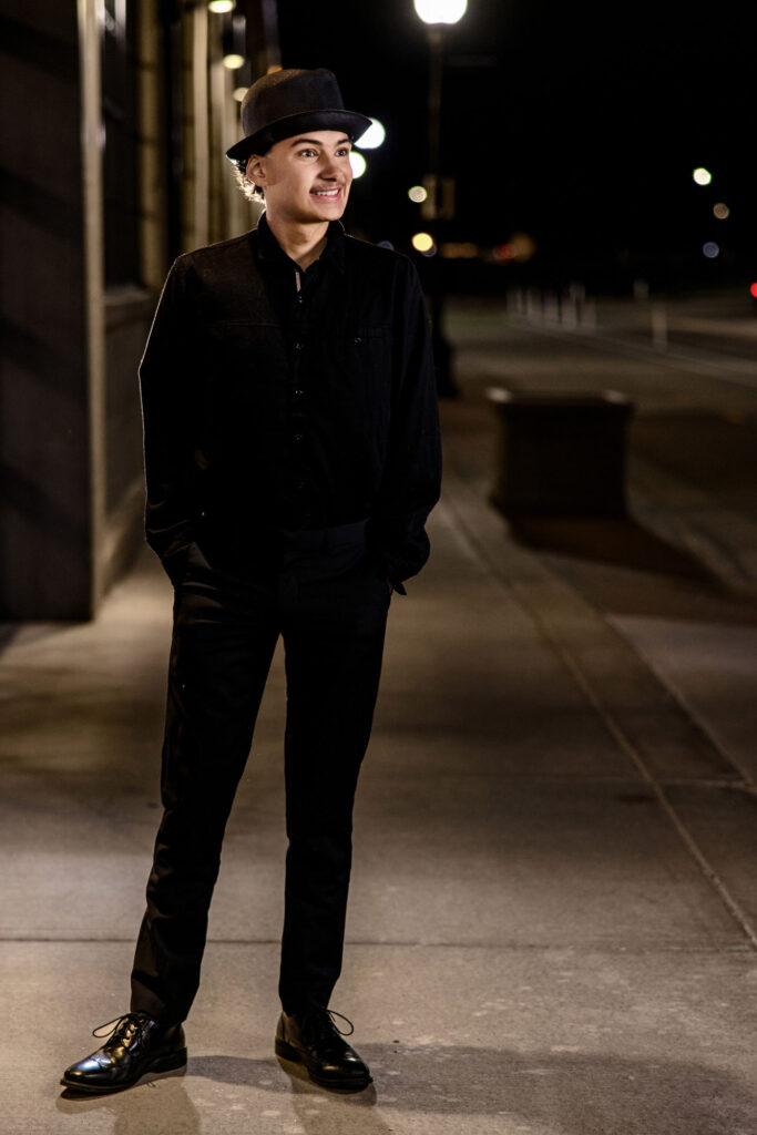 Young man in black jacket and cap standing on nighttime urban street, looking away with contemplative expression