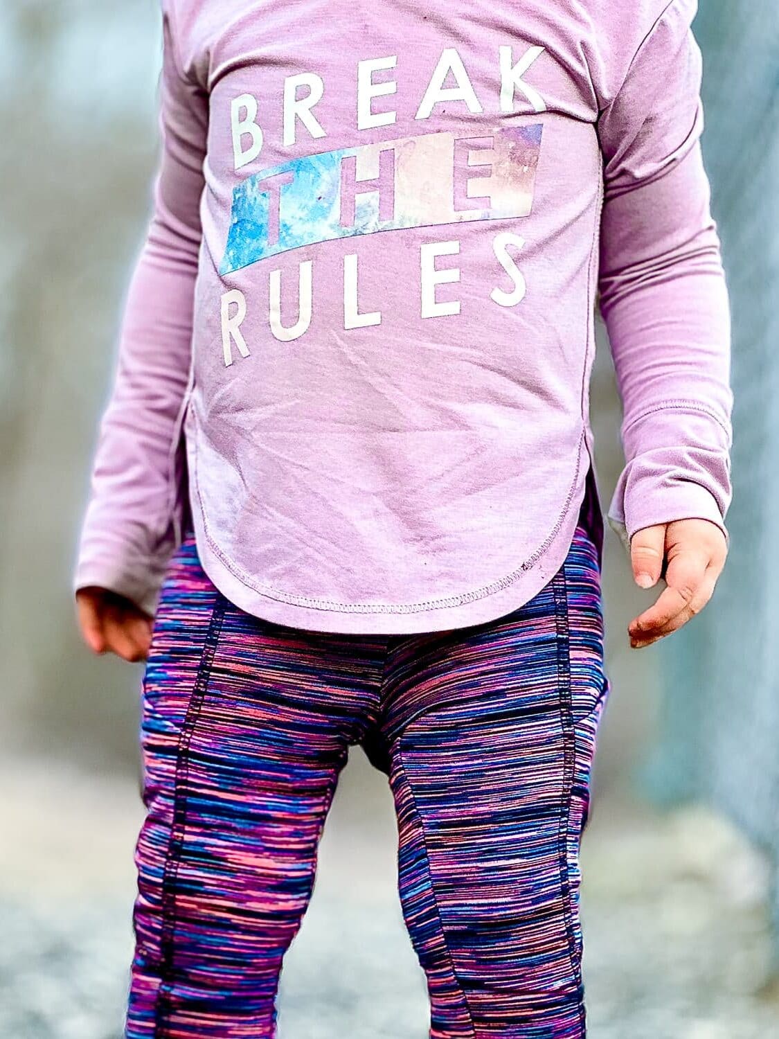 Child wearing pink ”Break the Rules” long-sleeve shirt and purple striped leggings, standing against neutral background