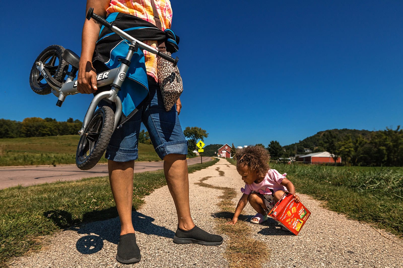 Adult holding a child's bicycle while a toddler explores a gravel path