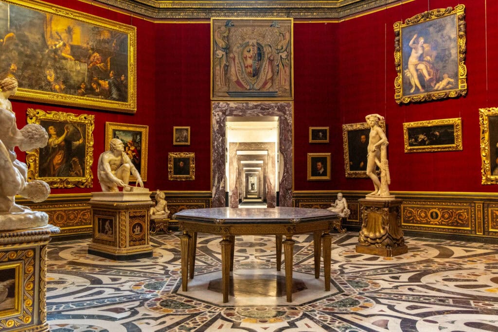 Ornate gallery room with crimson walls, gilded paintings, marble sculptures, and intricate inlaid floor in Florence, Italy