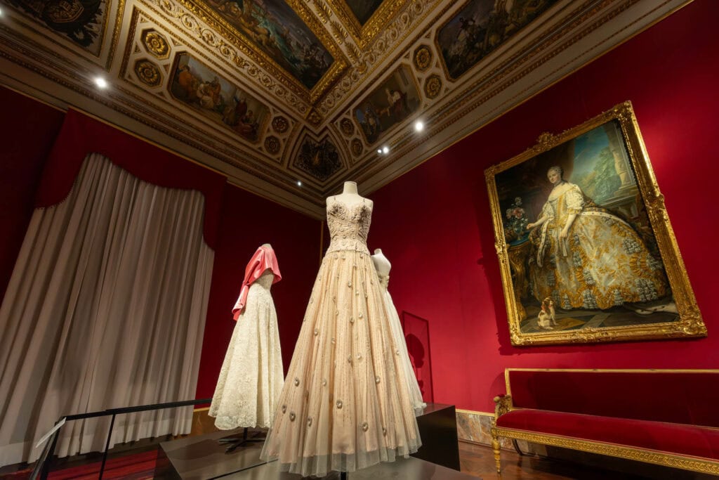 Two elegant cream gowns displayed on mannequins in ornate gallery with red walls and gilded ceiling, Florence