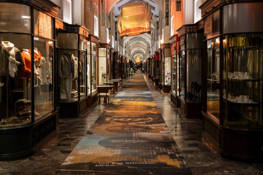 Illuminated European arcade with boutique storefronts, warm golden ceiling light, and patterned marble floor stretching into distance
