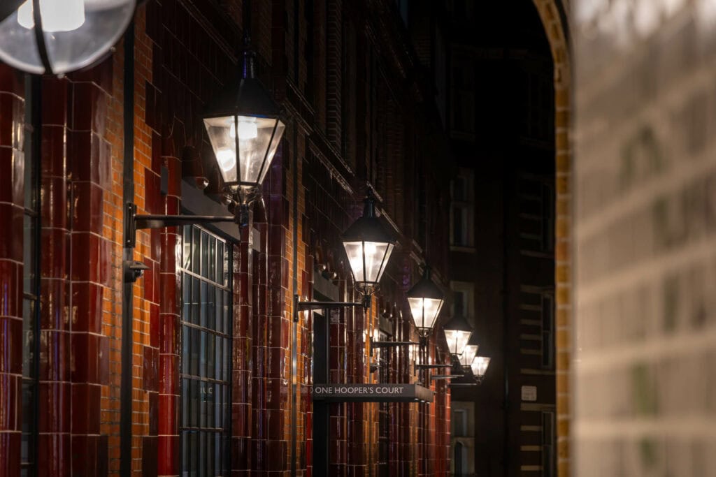 Narrow European alleyway lined with red brick buildings, lit by vintage street lamps at dusk, London or Florence