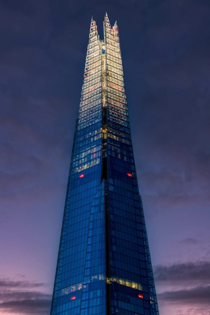 Illuminated skyscraper at dusk, its glass facade glowing blue and gold against a purple twilight sky, London