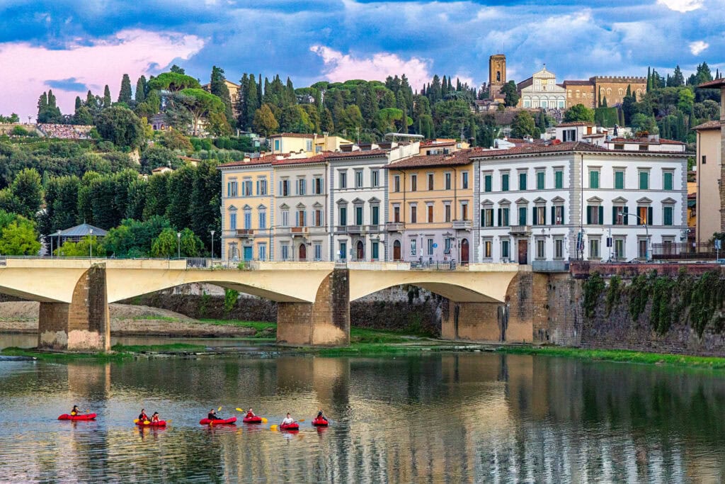 Row of red kayakers paddling on the Arno River below Renaissance palaces and stone bridge in Florence, Italy