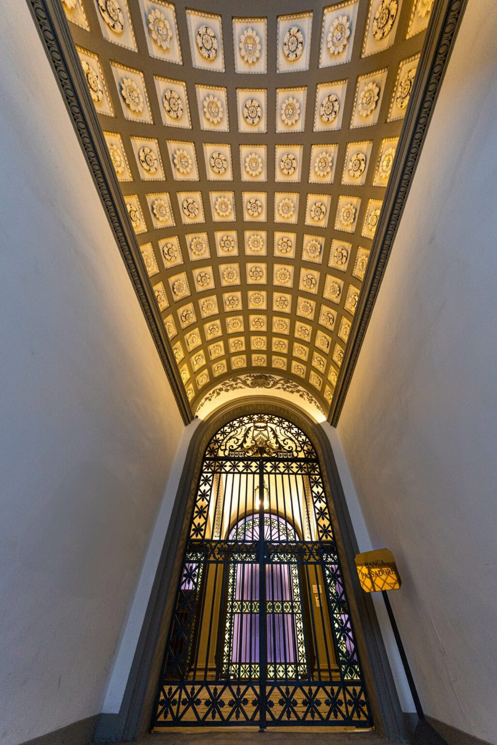 Ornate coffered ceiling with geometric patterns and glowing panels stretches toward decorative iron grille in historic London passage