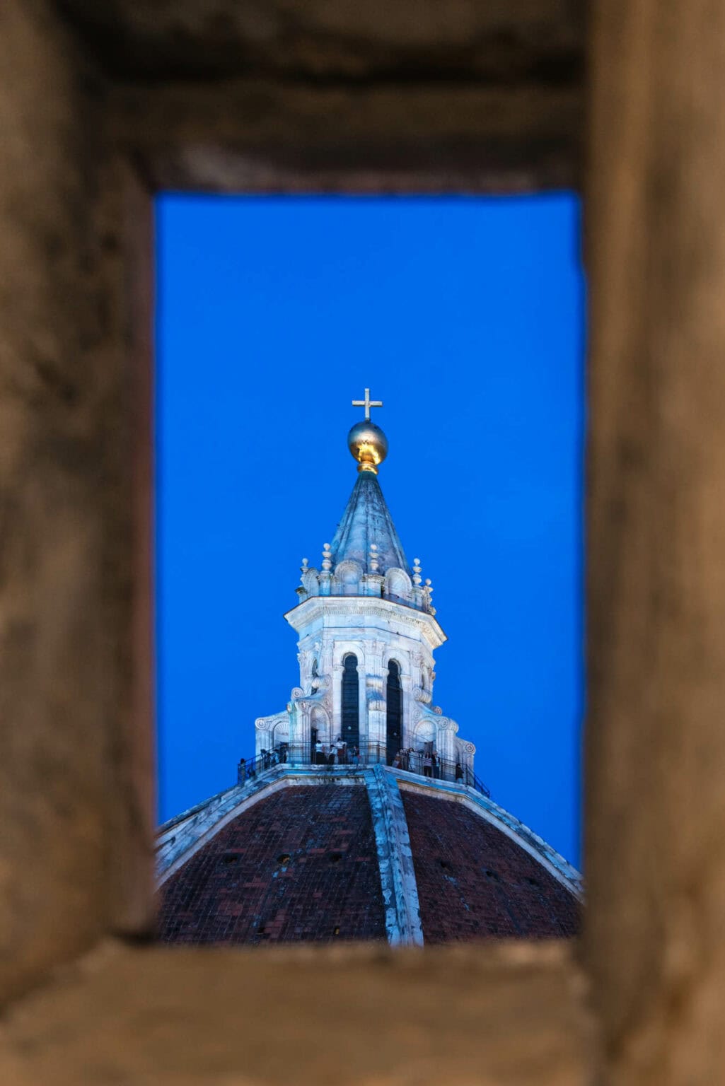 White dome and spire with golden cross framed through stone archway against brilliant blue sky, Florence Italy