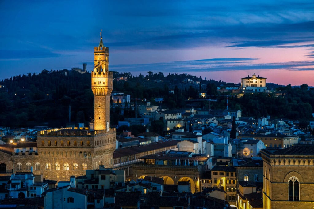 Palazzo Vecchio illuminated at twilight in Florence, Italy, with surrounding Renaissance architecture glowing against a blue evening sky
