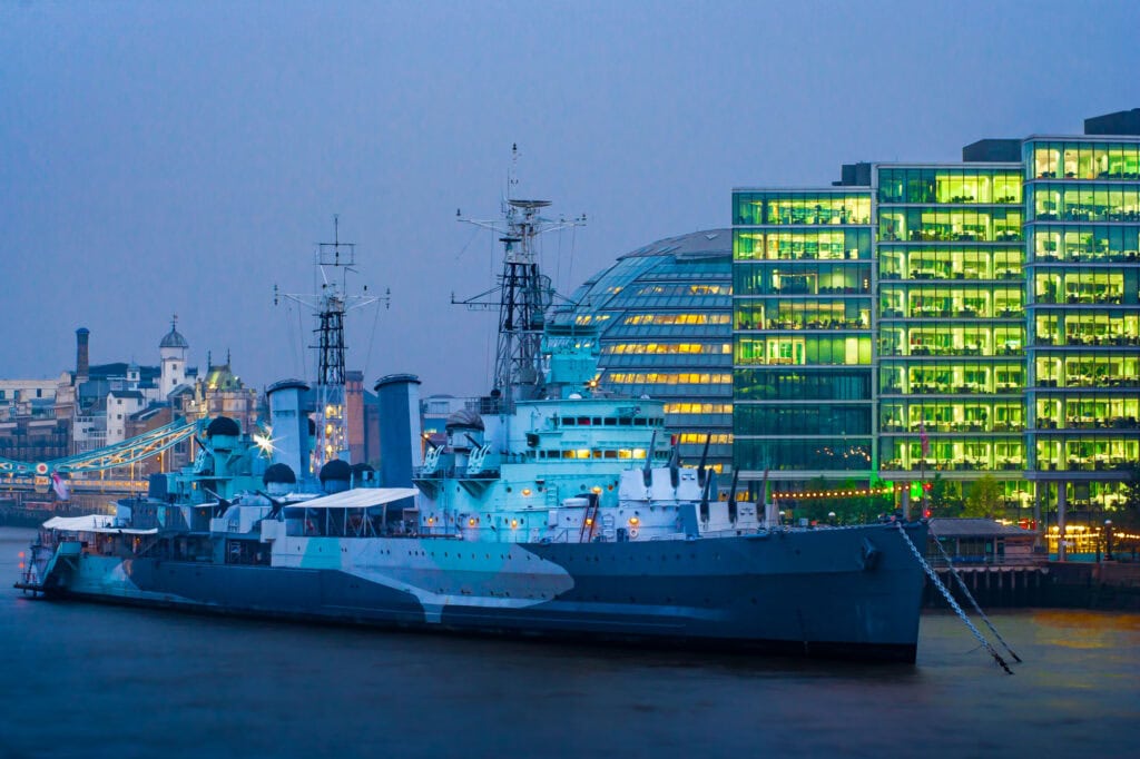 A blue research vessel, HMS Belfast, docked on the Thames at twilight among London’s illuminated buildings