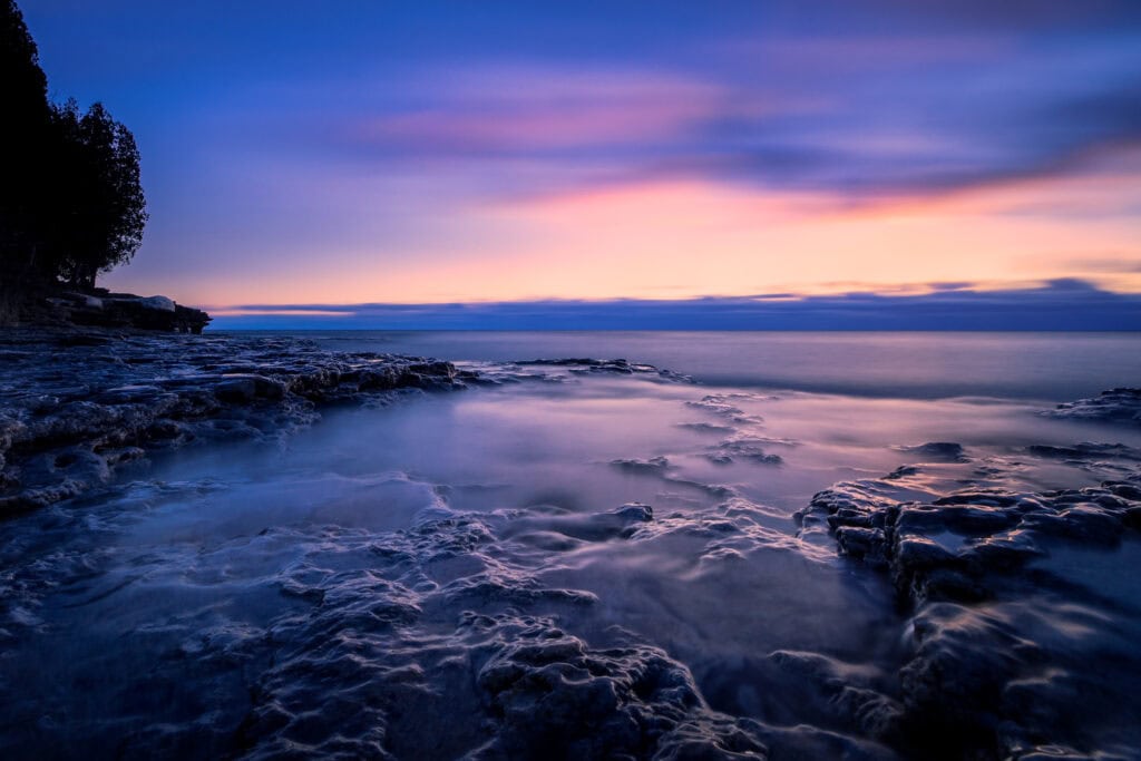 Cave Point at sunrise, waves breaking over the limestone shelves under a pink and violet sky