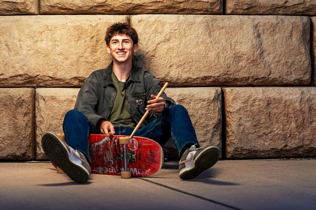 Emerson with his skateboard and drumsticks against a stone wall