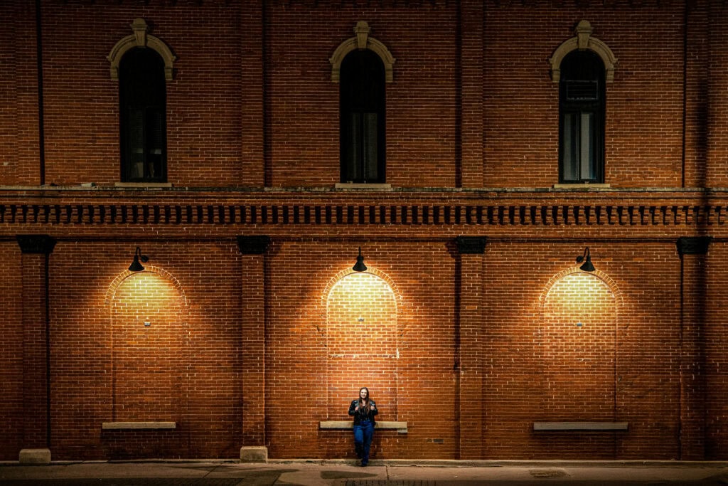 Claire on a bench beneath three lamps on a long brick wall at night, La Crosse