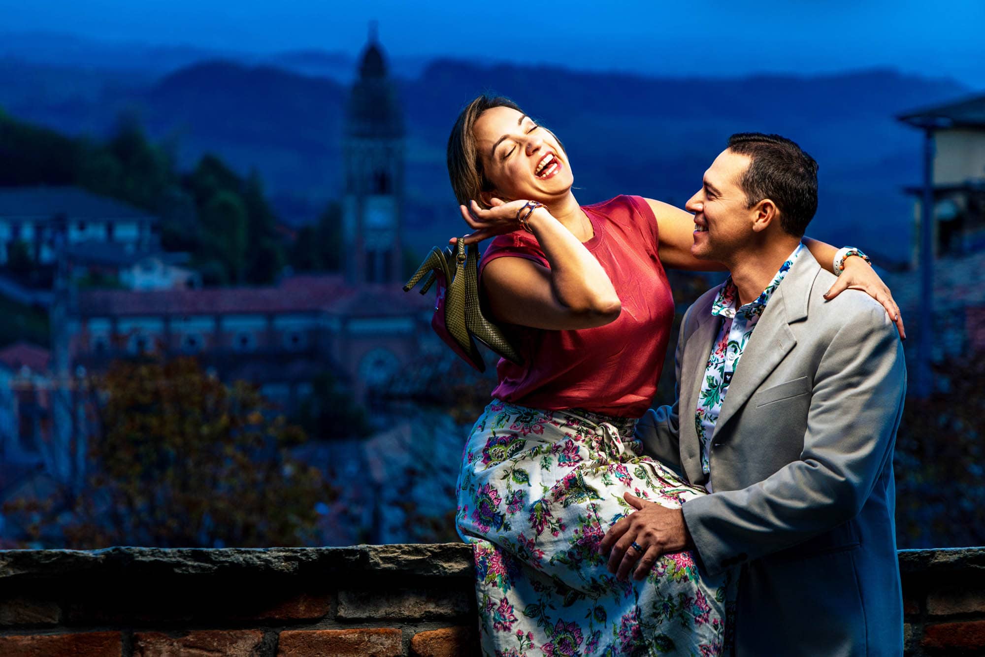 Couple laughing together on a stone terrace at dusk with a hillside town behind them