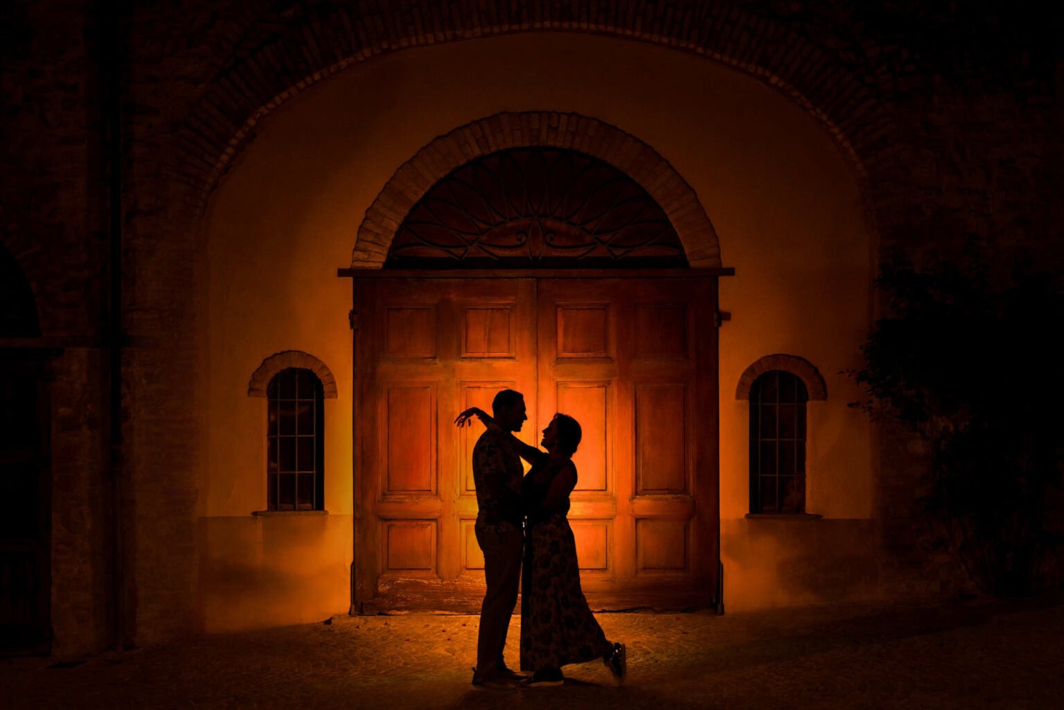 Couple embracing in silhouette in front of a glowing arched wooden door at night