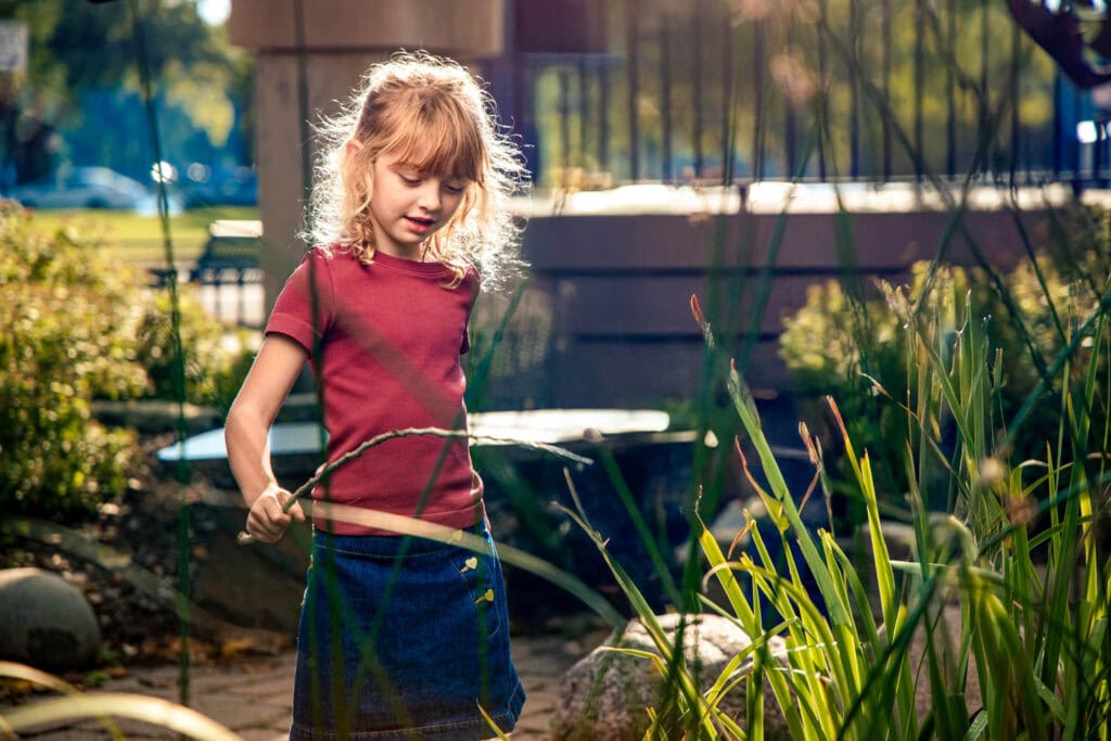 Young girl in maroon shirt exploring tall grasses in sunny garden setting during family portrait session
