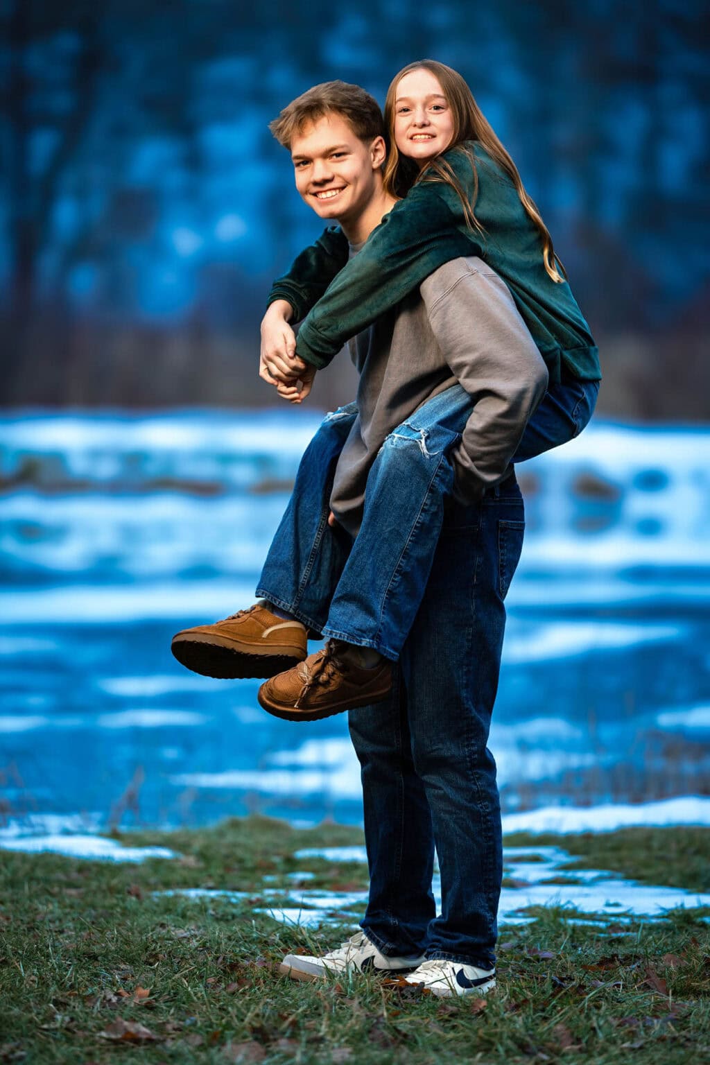 A brother carrying his sister piggyback beside the river at blue hour