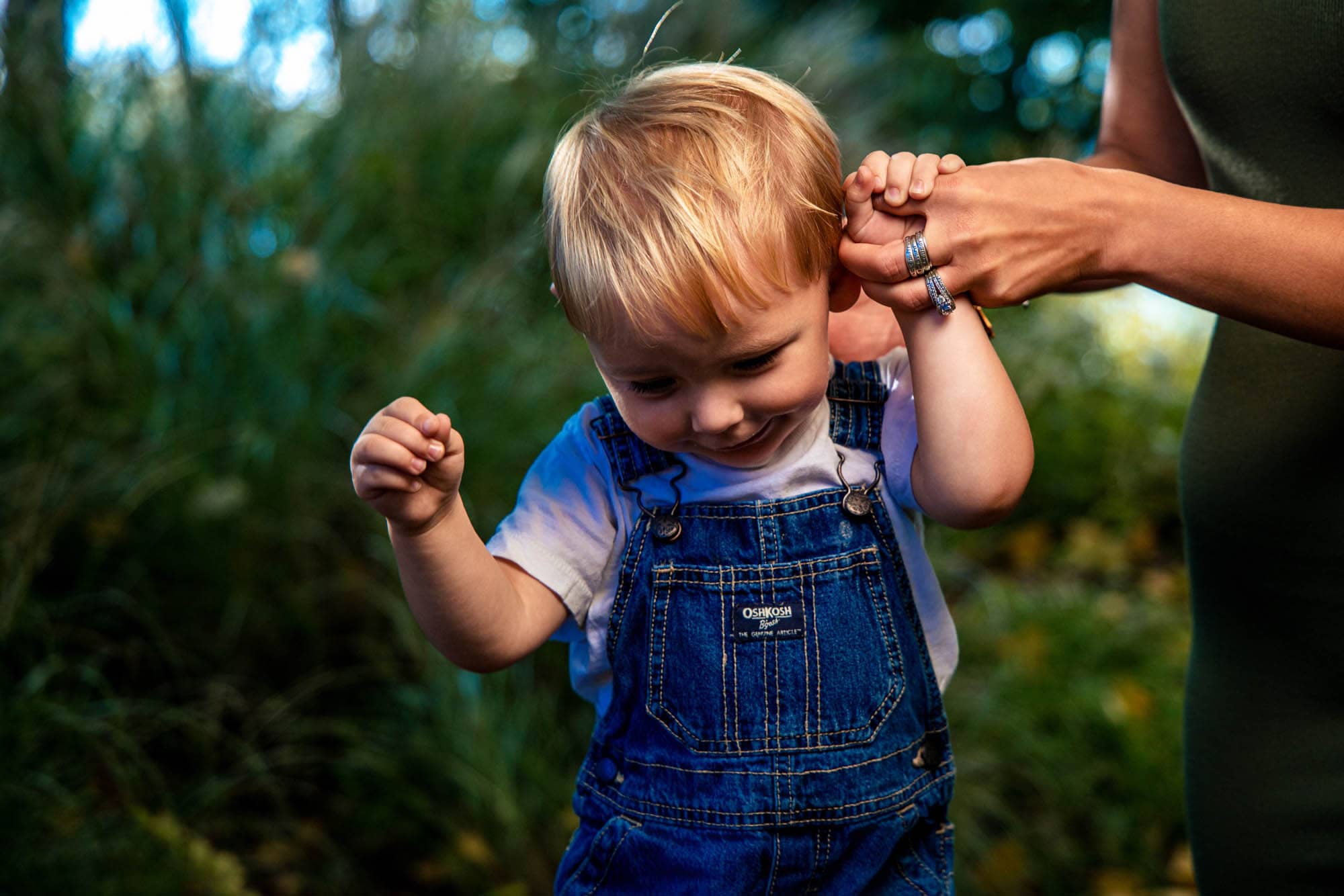Toddler in denim overalls holding his mom's hand while learning to walk in the garden