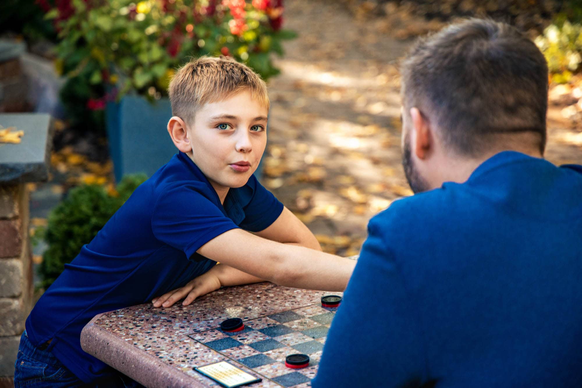 Boy looks back over his shoulder while playing checkers with his dad at an outdoor table