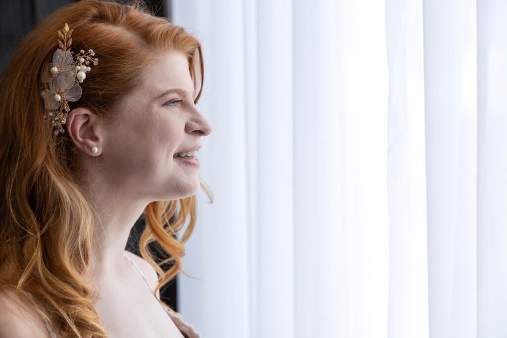 Redheaded bride in profile smiling softly, gold floral hair piece catching the window light
