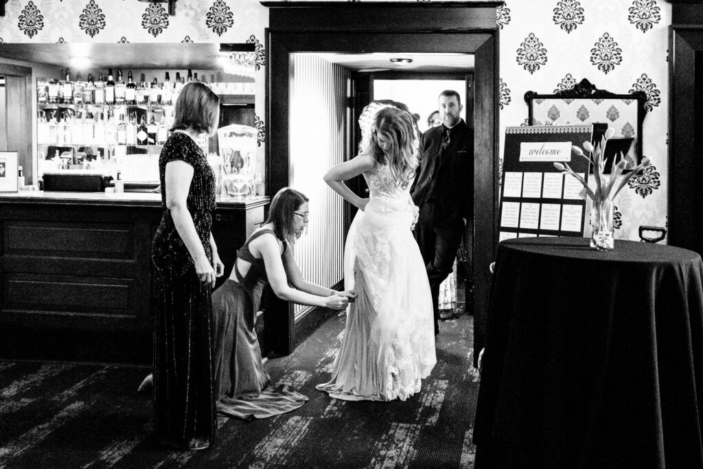 Bridesmaid kneeling to straighten the bride's gown hem inside a vintage venue doorway