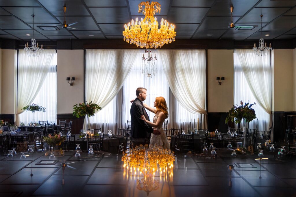 Couple standing face to face on an empty dance floor beneath amber chandeliers and draped white curtains