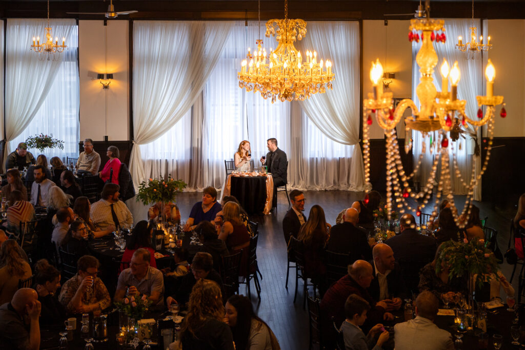 Bride and groom at their sweetheart table address a full wedding reception hall glowing with gold chandeliers in La Crosse, W