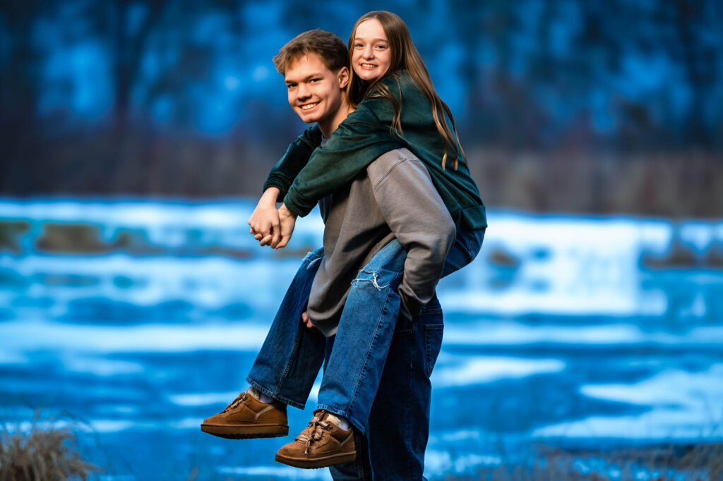 Teen boy and younger girl smiling together on a grassy hilltop with golden fall foliage behind them in La Crosse, WI