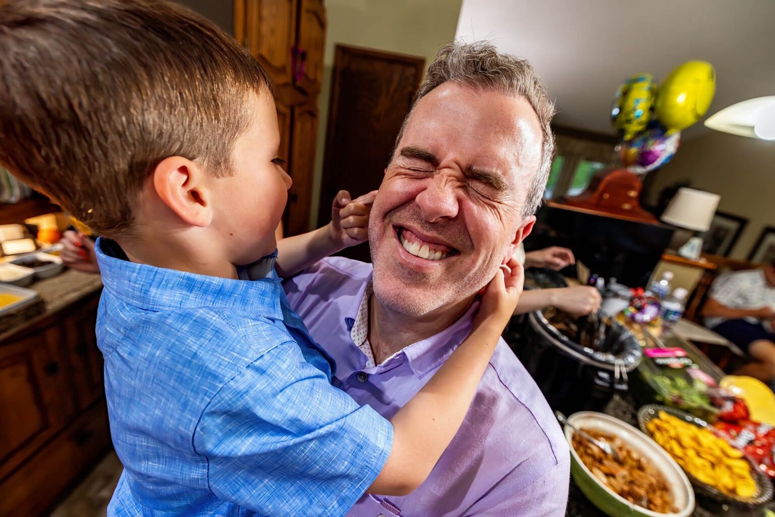 A young boy squishes his laughing dad’s face in the kitchen during a documentary family session at home