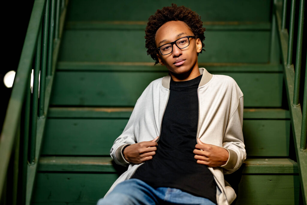 High school senior in glasses and open white jacket holding lapels, direct confident gaze, green staircase backdrop