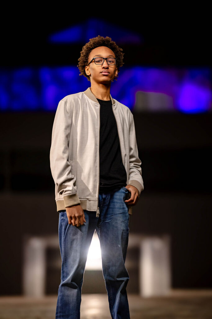 High school senior standing full-length on downtown street at night, blue architectural glow behind, dramatic flash from…