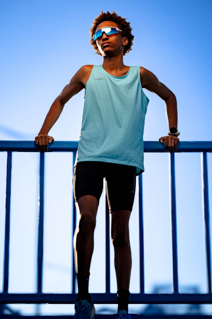 Teen boy in teal tank and mirrored sunglasses stands with arms spread on the Cass Street Bridge railing, photographed from…