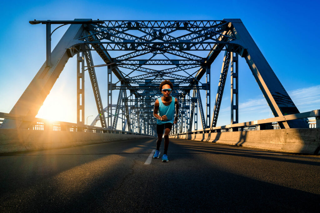 Young male runner sprints toward camera on Cass Street Bridge at sunrise with golden sun flare through steel trusses, La…