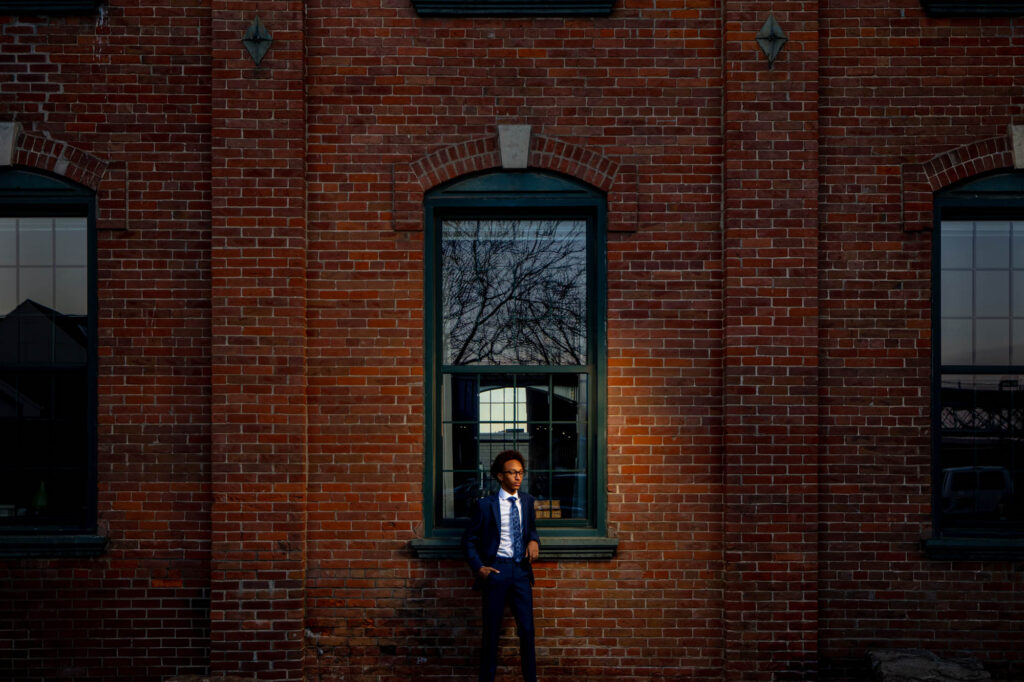 High school senior in navy suit leaning against a grand red brick building in downtown La Crosse, a dramatic shaft of…