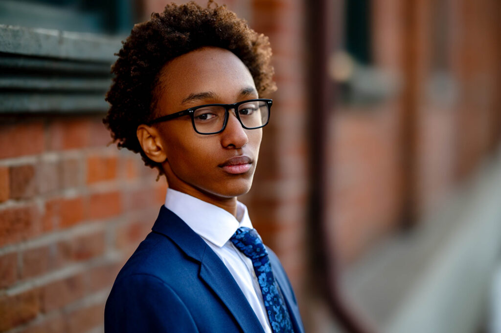 Close-up portrait of high school senior in navy suit and glasses with a composed, faraway gaze against red brick, La Crosse…
